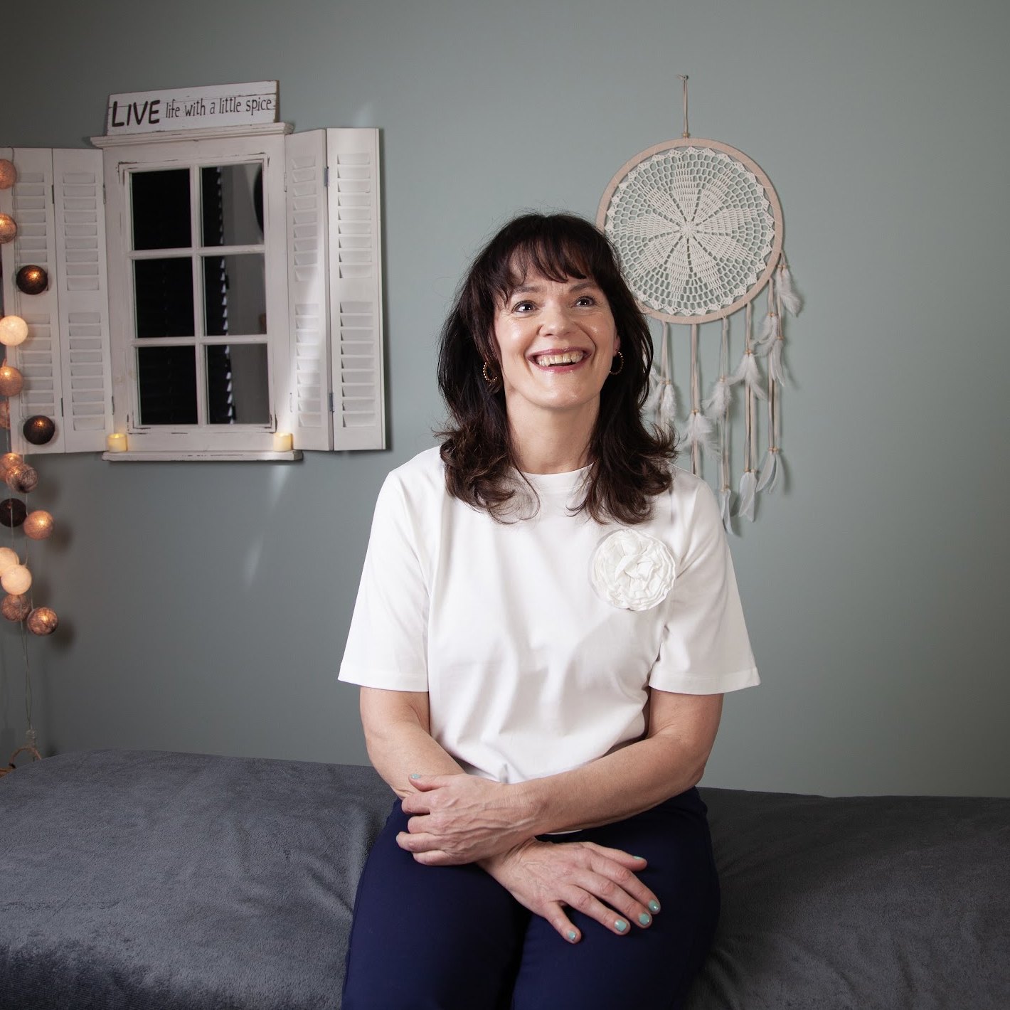 Adriënne zit lachend op haar massagetafel, met een dromenvanger aan de muur achter haar